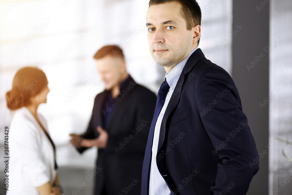 Portrait of a self-confident middle aged businessman in a blue suit, standing in a sunny modern office with his colleagues at the background. Concept of business success Portrait of a self-confident middle aged businessman in a blue suit, standing in a sunny modern office with his colleagues at the background. Concept of business success