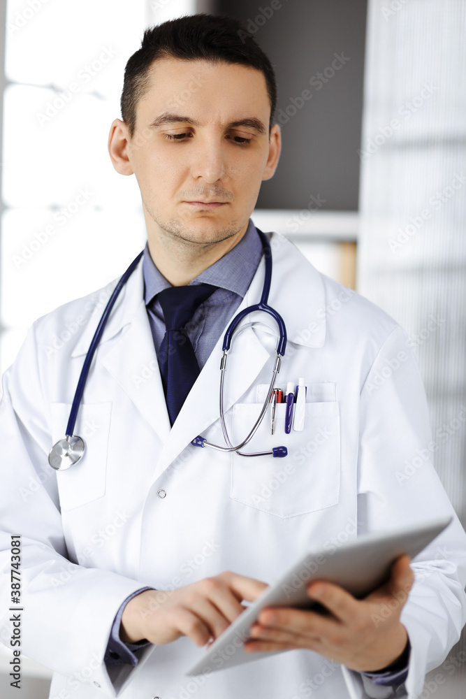 Friendly male doctor using tablet computer in clinic near his working place. Perfect medical service in hospital. Medicine and healthcare concept Friendly male doctor using tablet computer in clinic near his working place. Perfect medical service in hospital. Medicine and healthcare concept