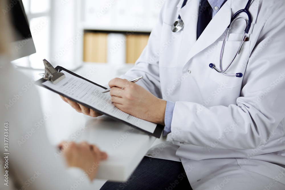 Unknown male doctor and patient woman discussing current health examination while sitting in clinic and using clipboard. Perfect medical service in hospital. Medicine and healthcare concept Unknown male doctor and patient woman discussing current health examination while sitting in clinic and using clipboard. Perfect medical service in hospital. Medicine and healthcare concept