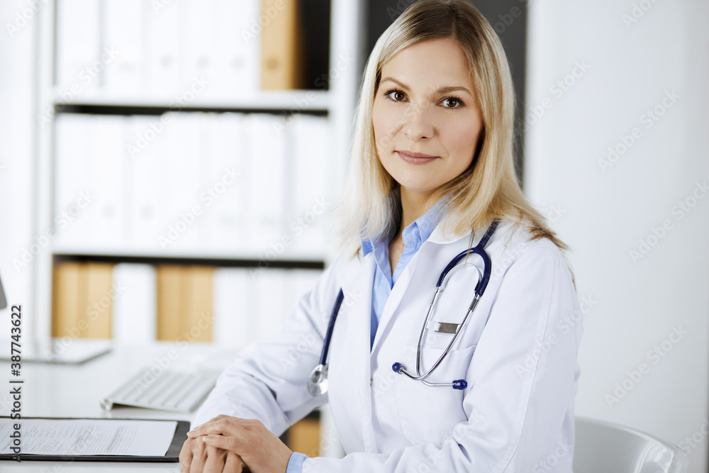 Friendly female doctor sitting in clinic at her working place. Portrait of cheerful smiling physician. Perfect medical service in hospital. Medicine and healthcare concept Friendly female doctor sitting in clinic at her working place. Portrait of cheerful smiling physician. Perfect medical service in hospital. Medicine and healthcare concept