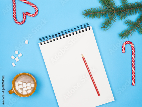 White note for christmas wish list or goals and resolutions in new year red pencil, candy cane cocoa, christmas treeand clay mug with hote cocoa and marshmallow on blue winter background	 photo