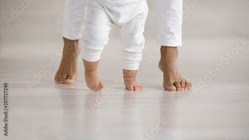 Crop close up of little baby infant learn walking at home wooden floor holding mom hands. Cute small toddler child make first steps with mom love, care and support. Childcare, motherhood concept.