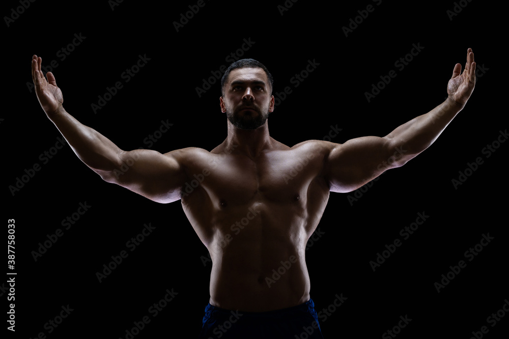 Portrait of a bodybuilder standing isolated on black background in a shadow with raised hands to show off his muscles