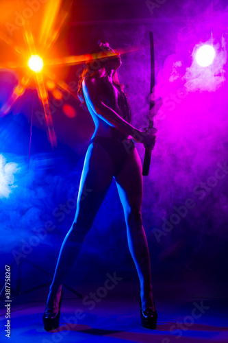 Beautiful slim sexy girl wearing lingerie and high heels posing holding katana sword in her hand in the rays of light in a colorful smoke. Artistic, conceptual, silhouette and advertising design © Алексей Торбеев