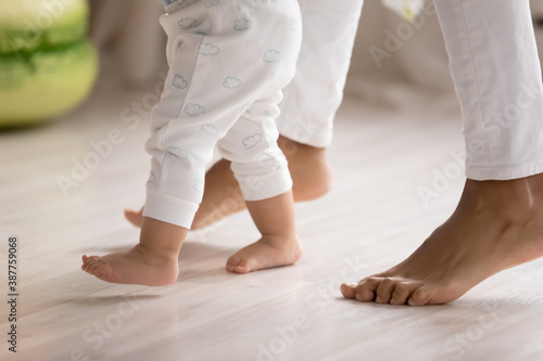 Crop close up of loving mother hold little biracial child hands learn walking at home on wooden floor. Small cute ethnic baby toddler make first steps with mom support and care. Childcare concept.