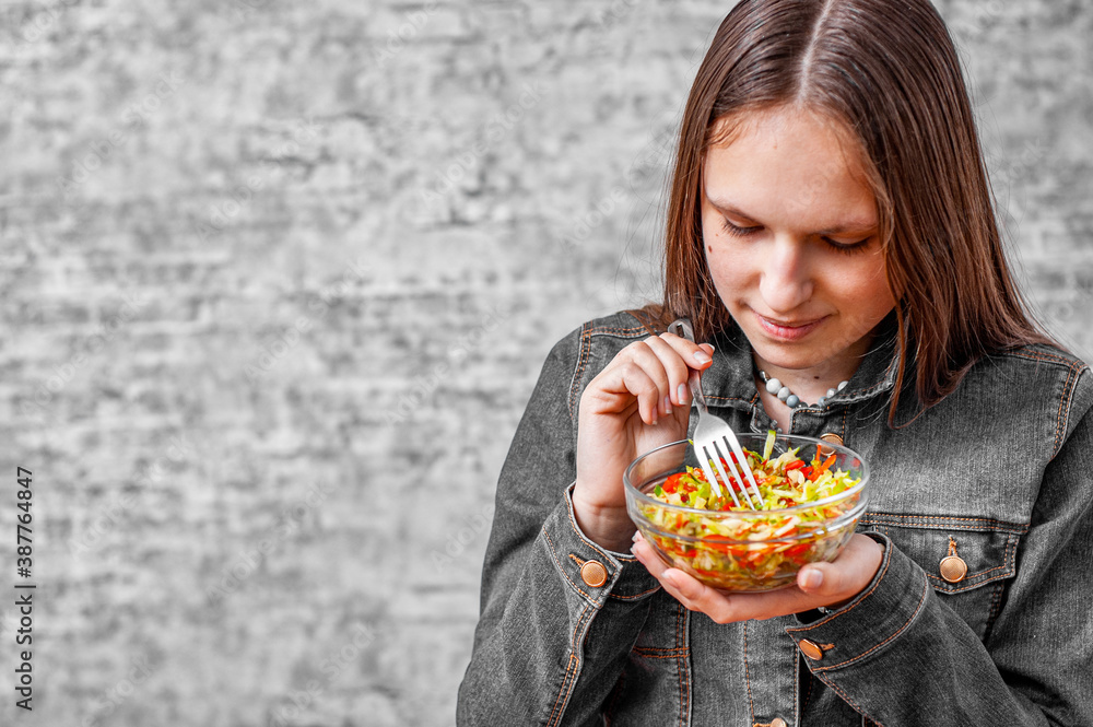 portrait of young teenager brunette girl with long hair eating green vegetables salad on gray wall background with copy space.