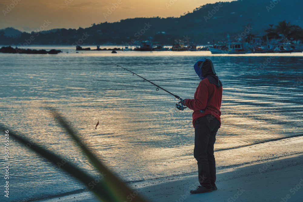 Adult Thai woman in protective suit and mask fishing on the beach with spinning reel at sunset 