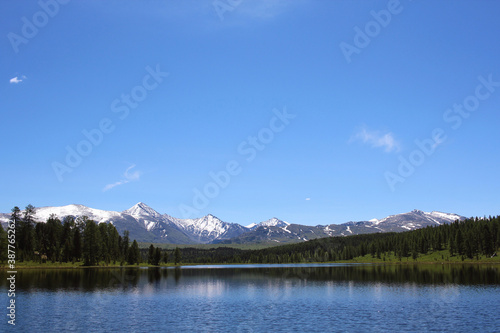 The snow-covered mountain range and green forest are reflected in the crystal blue waters of the lake. Pastoral meditative landscape. Mountain Lake. Wildlife. Travel across Russia