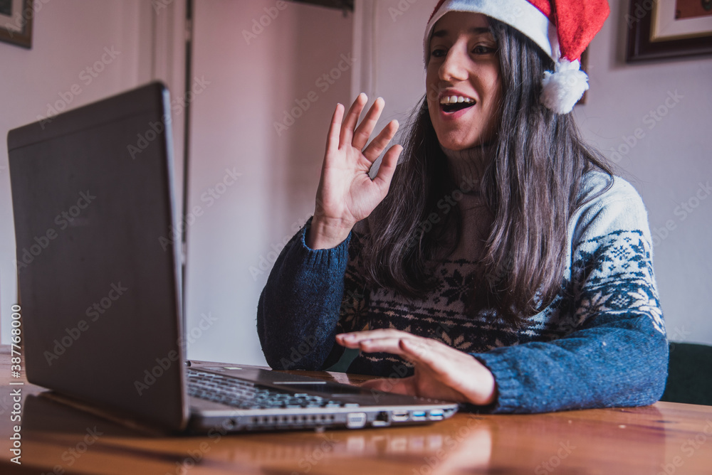 Una chica joven haciendo una video llamada saluda a su ordenador portátil  mientras lleva puesto un gorro de Santa Claus y sujeta su teléfono móvil  foto de Stock | Adobe Stock