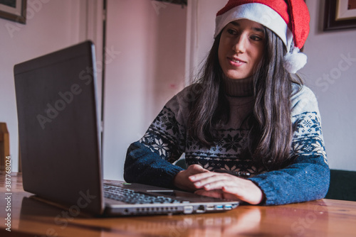 Una chica joven sonríe mientras trabaja con su ordenador portátil mientras lleva puesto un gorro de Santa Claus y un jersey de Navidad