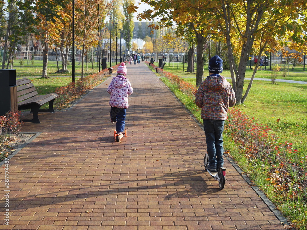 Fototapeta premium Happy children ride a scooter in the park. A boy and a girl rest in the autumn outdoors. Back view.