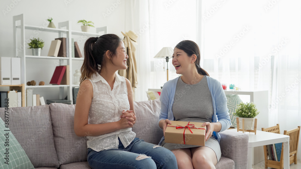 Happy asian japanese pregnant woman with female friend sitting on sofa and celebrating baby shower in modern bright apartment.