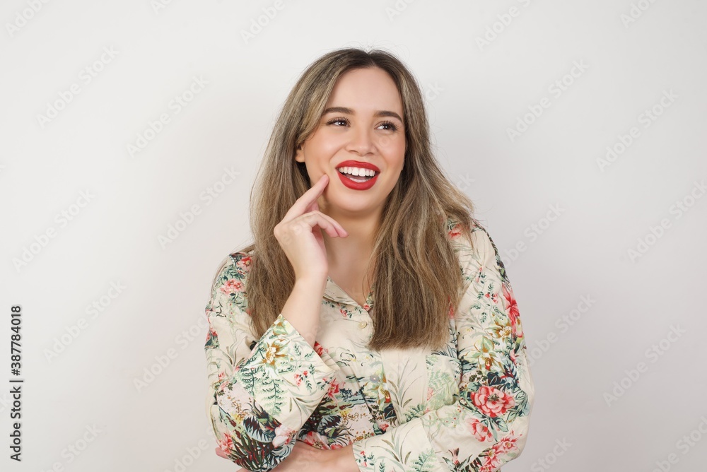 Dreamy female with thoughtful expression, looks to the camera, keeps hand near face, bitting a finger thinks about something pleasant, poses against gray wall. Dreamy female with thoughtful expression, looks to the camera, keeps hand near face, bitting a finger thinks about something pleasant, poses against gray wall.