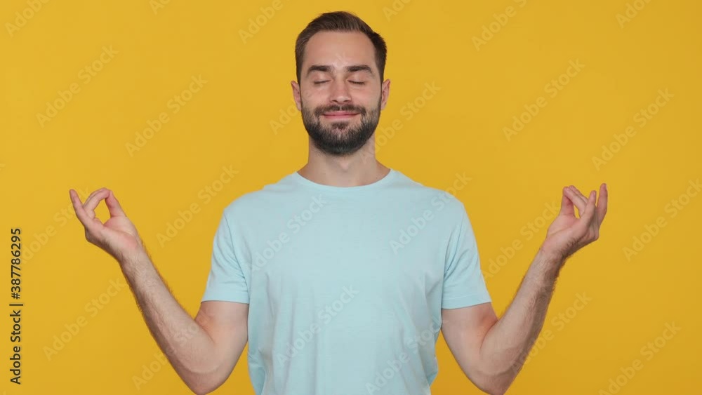 Handsome sporty bearded young man 20s in basic casual blue t-shirt hold hands in yoga gesture relaxing meditating trying to calm down isolated on yellow wall background studio People lifestyle concept Handsome sporty bearded young man 20s in basic casual blue t-shirt hold hands in yoga gesture relaxing meditating trying to calm down isolated on yellow wall background studio People lifestyle concept