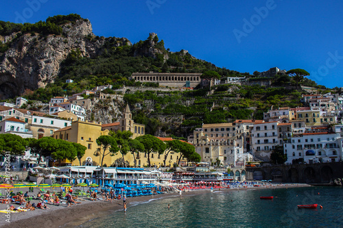 View of the beach with its bathers from the town of Amalfi from the jetty with the sea, boats and colorful houses on the slopes of the Amalfi coast in the province of Salerno, Campania, Italy.