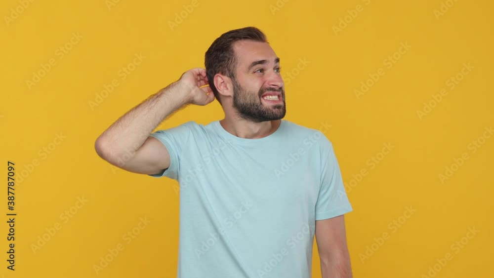 Confused thoughtful fun bearded young man 20s in basic casual blue t-shirt looking around thinking scratches at temple gesture isolated on yellow background studio. People emotions lifestyle concept Confused thoughtful fun bearded young man 20s in basic casual blue t-shirt looking around thinking scratches at temple gesture isolated on yellow background studio. People emotions lifestyle concept