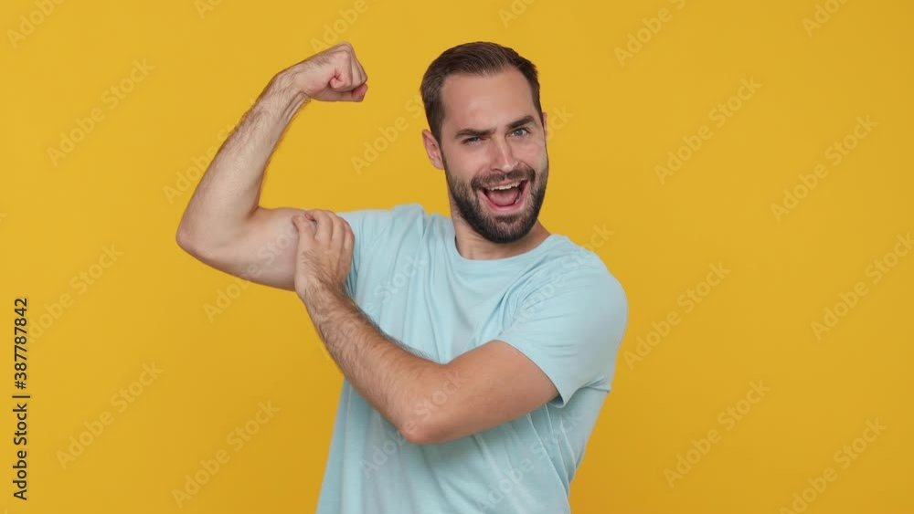 Young strong man 20s years old in basic casual blue t-shirt isolated on yellow background studio. People sincere emotions lifestyle concept. Looking camera charming smile showing biceps muscles hand Young strong man 20s years old in basic casual blue t-shirt isolated on yellow background studio. People sincere emotions lifestyle concept. Looking camera charming smile showing biceps muscles hand