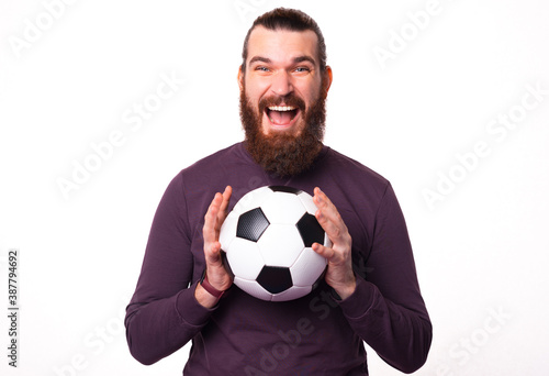 Young bearded man is looking excited at the camera and holding a soccer ball .
