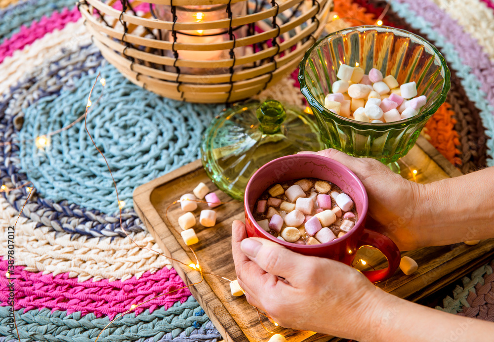 Hands holding red cup of coco with pastel color marshmallows, wire and bamboo lantern with candle on colorful crochet rug from repurposed T-shirts. Cozy autumn winter set.