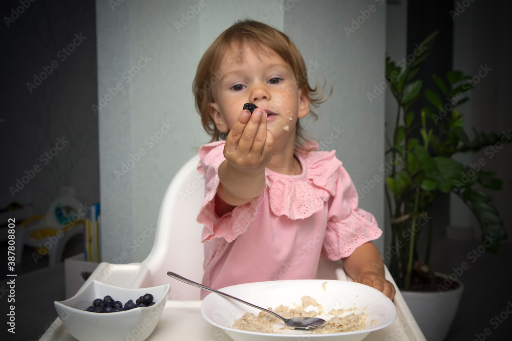 Happy, smiling child eating oatmeal with berries. The concept of a healthy breakfast for children, porridge with blueberries on baby chair Happy, smiling child eating oatmeal with berries. The concept of a healthy breakfast for children, porridge with blueberries on baby chair