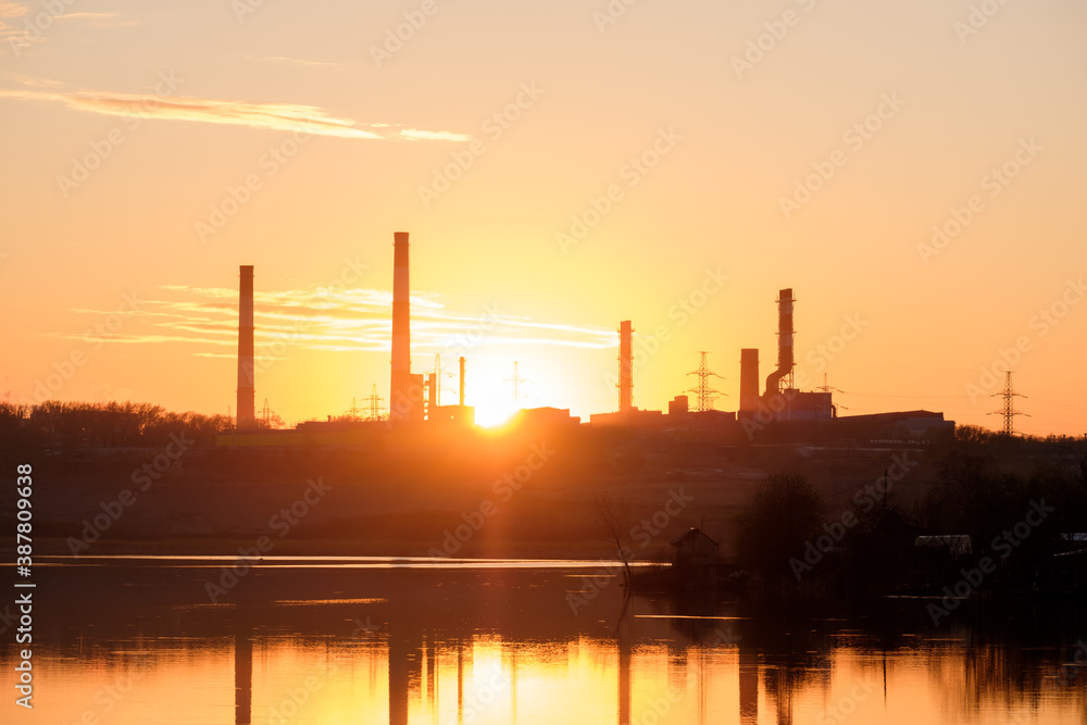 rays of the sun through the factory chimneys during sunset on the shore of the reservoir