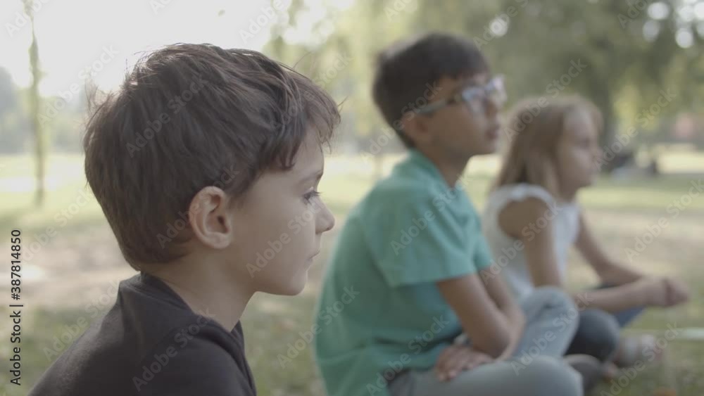 Side portrait of lovely Caucasian boy looking away. Multiethnic concentrated children sitting on ground cross-legged. Selective focus, Static camera. Childhood, weekend and summer vacation concept Side portrait of lovely Caucasian boy looking away. Multiethnic concentrated children sitting on ground cross-legged. Selective focus, Static camera. Childhood, weekend and summer vacation concept