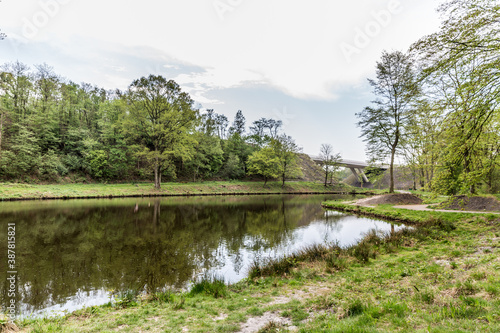 River with calm water and mirror reflection with green grass and lush trees on the shore with a bridge in the background  cloudy day with cloud covered sky in South Limburg  Netherlands