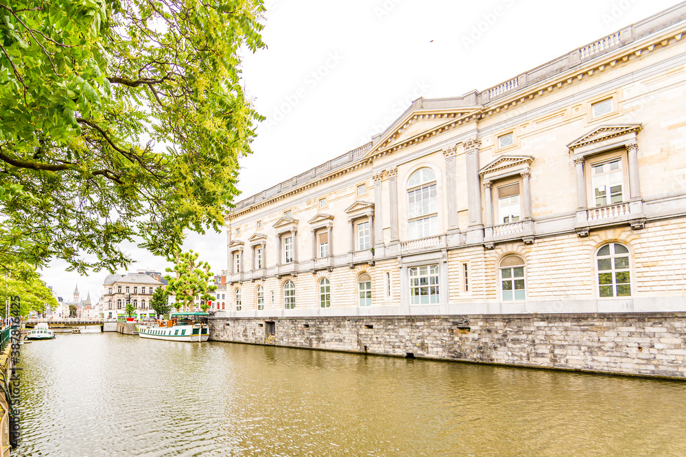 The Palace of Justice in Ghent next the Leie River with anchored boats and trees along the river, a relaxed spring day with cloudy sky in Belgium