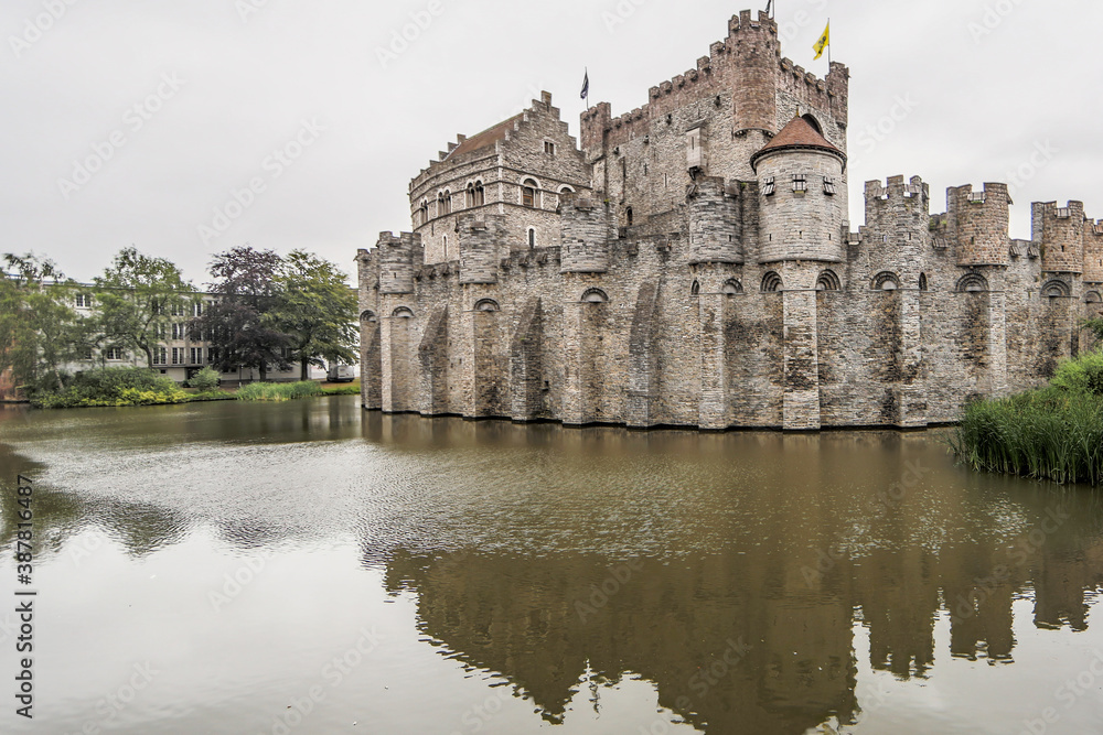 Canal with calm waters surrounding the medieval castle of Gravensteen (Castle of the Counts), reflection and light breeze on the water surface, cloudy day with a gray sky in Ghent, Belgium