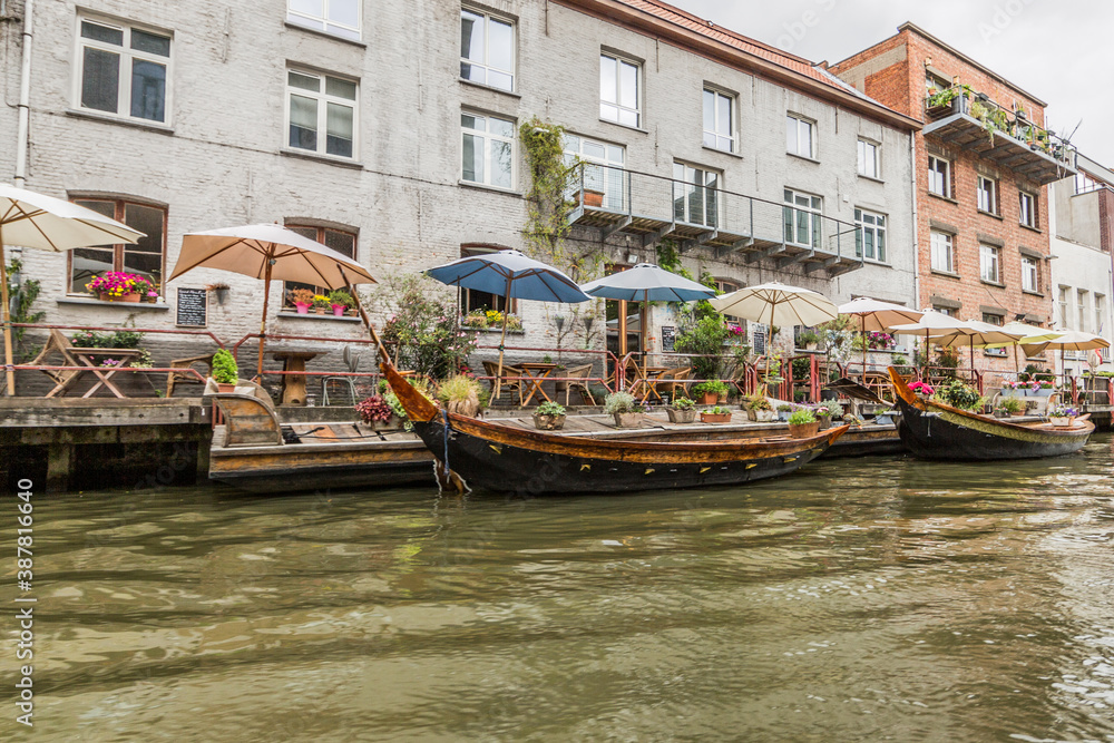 Picturesque terrace with its colorful umbrellas, wooden tables and chairs, flowers next to a building with many windows, boats anchored on the canal pier, cloudy day in the city of Ghent, Belgium