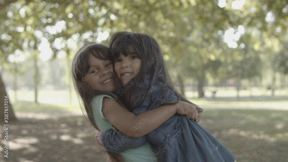 Cute Latin girls hugging each other and smiling. Lovely cheerful kids standing in city park, laughing, embracing and holding hands. Static camera. Childhood, friendship and summer activity concept Cute Latin girls hugging each other and smiling. Lovely cheerful kids standing in city park, laughing, embracing and holding hands. Static camera. Childhood, friendship and summer activity concept