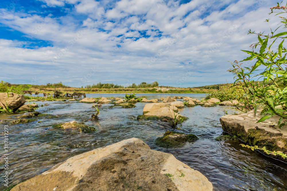 Fototapeta premium Clean water flowing between the rocks in a stony stream until it empties into the Maas river among wild vegetation, sunny day, with a blue sky in Elsloo and Meers South Limburg, Netherlands Holland