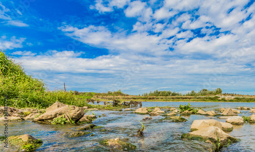 Clean water flowing into the Maas river through a stony stream among wild vegetation with few trees in the background, sunny day with a blue sky in Elsloo and Meers South Limburg, the Netherlands