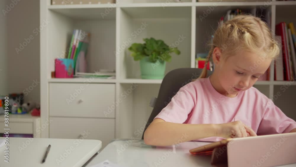 Tracking medium shot of cute elementary school girl using digital tablet for learning sitting at desk at home Tracking medium shot of cute elementary school girl using digital tablet for learning sitting at desk at home