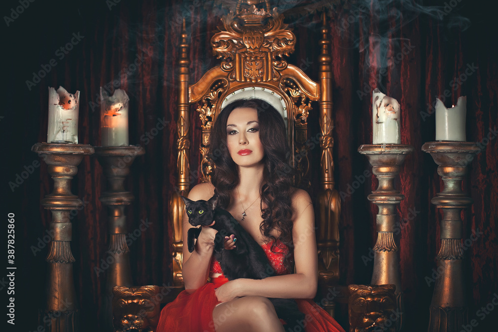 A brunette girl in a red dress sitting on a throne with a black cat in her arms against a background of candelabra with candles. Halloween theme, witch cosplay.