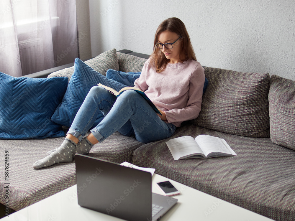 A young girl is sitting on the couch and reading a book. Female student studying at home. There is a laptop and a book on the table. A young girl is sitting on the couch and reading a book. Female student studying at home. There is a laptop and a book on the table.