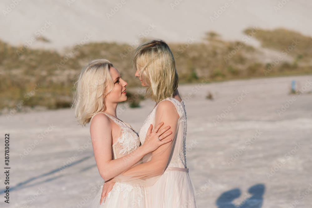 Beautiful lesbian couple walking on sand along river bank on their wedding day Beautiful lesbian couple walking on sand along river bank on their wedding day