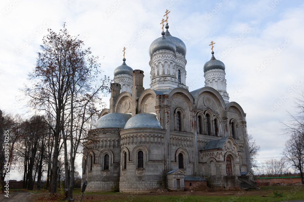 White stone church with silver domes in russian village Sergeevo in sunny autumn day with green grass, naked trees, blue sky