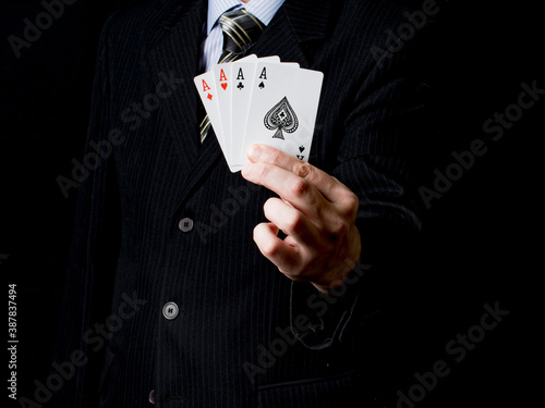 A man in a black suit and a tie holds 4 aces in his hand with black background. Poker cards. Play in poker and gambling games. Use for gambling sites, articles, advertising, gifts.
