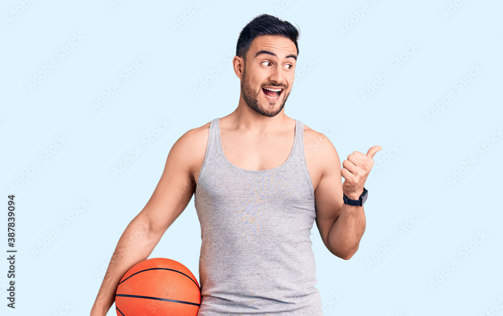 Young handsome man holding basketball ball pointing thumb up to the side smiling happy with open mouth Young handsome man holding basketball ball pointing thumb up to the side smiling happy with open mouth