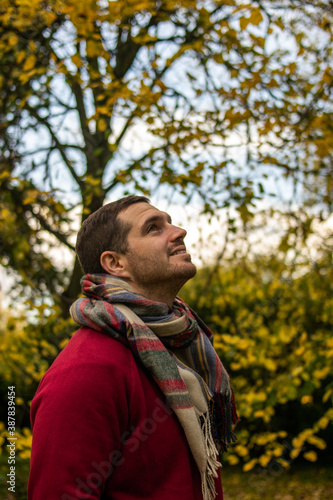 Photo of a young and attractive man wearing a scarf looking to the trees that surround him, enjoying a beautiful autumn day in nature