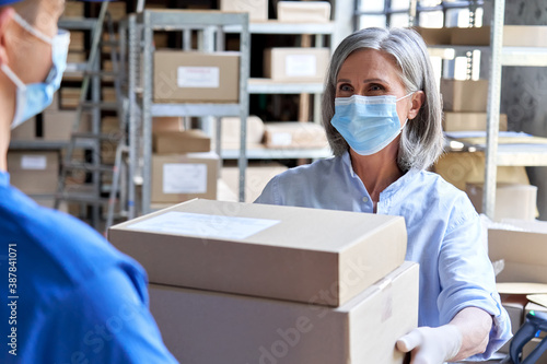 Female manager worker wearing face mask holding fast drop shipping safe delivery packages giving parcels shipment boxes to male courier taking ecommerce orders to deliver in warehouse storage.