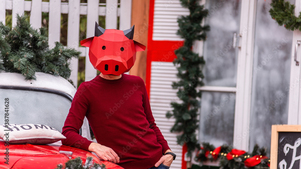 Obraz premium A young man in a bull ox costume is a symbol of the new year against the background of a red car, a festive Christmas tree, decorated with garlands of porch lights with a wreath. Copy space, template