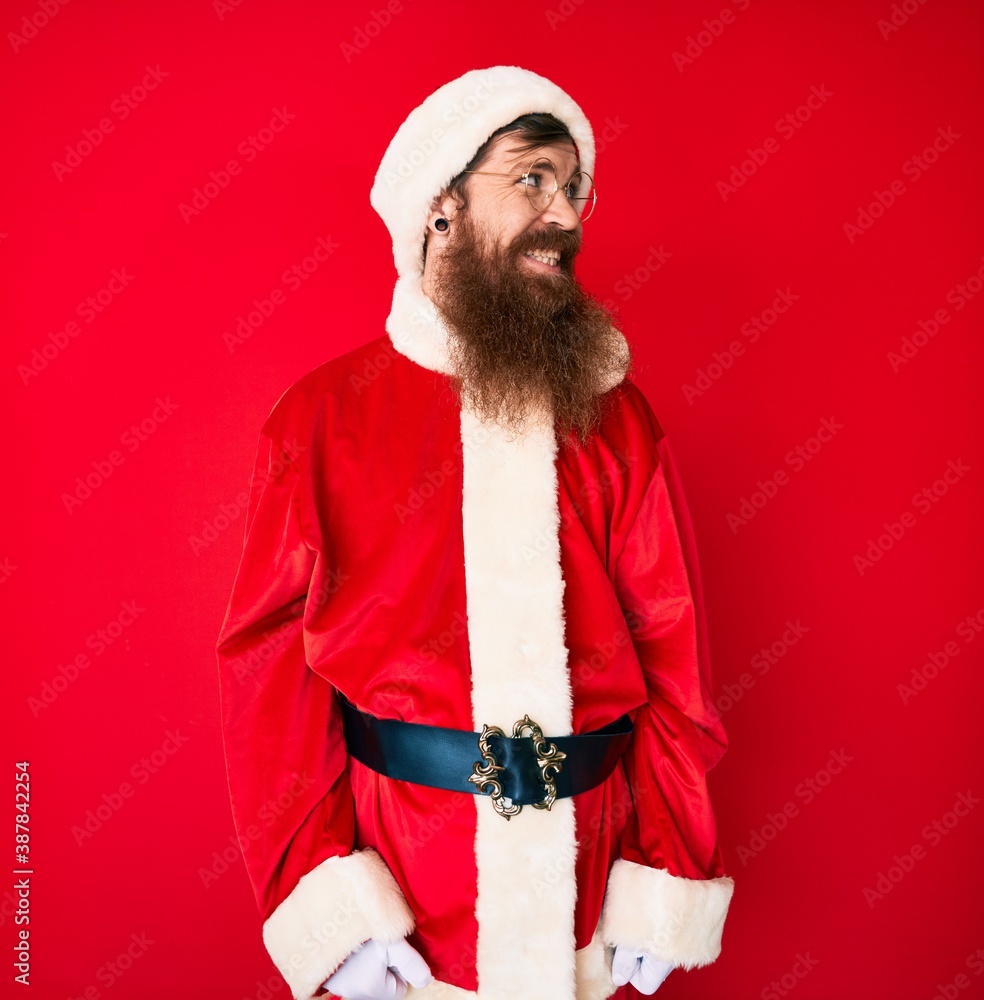 Handsome young red head man with long beard wearing santa claus costume looking away to side with smile on face, natural expression. laughing confident. Handsome young red head man with long beard wearing santa claus costume looking away to side with smile on face, natural expression. laughing confident.