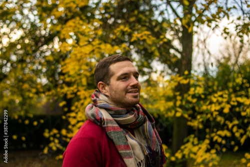 Photo of a young and attractive man wearing a scarf looking to the trees that surround him, enjoying a beautiful autumn day in nature