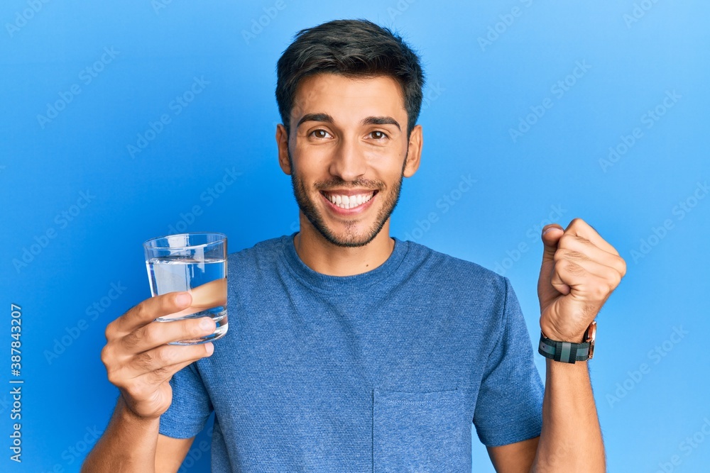 Young handsome man drinking glass of water screaming proud, celebrating victory and success very excited with raised arm Young handsome man drinking glass of water screaming proud, celebrating victory and success very excited with raised arm