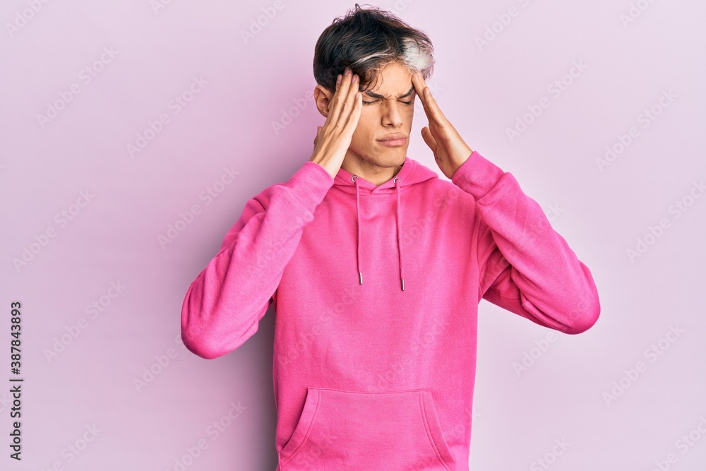 Young hispanic man wearing casual pink sweatshirt with hand on head, headache because stress. suffering migraine. Young hispanic man wearing casual pink sweatshirt with hand on head, headache because stress. suffering migraine.