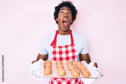 Handsome african american man with afro hair wearing baker uniform holding homemade bread angry and mad screaming frustrated and furious, shouting with anger looking up.
