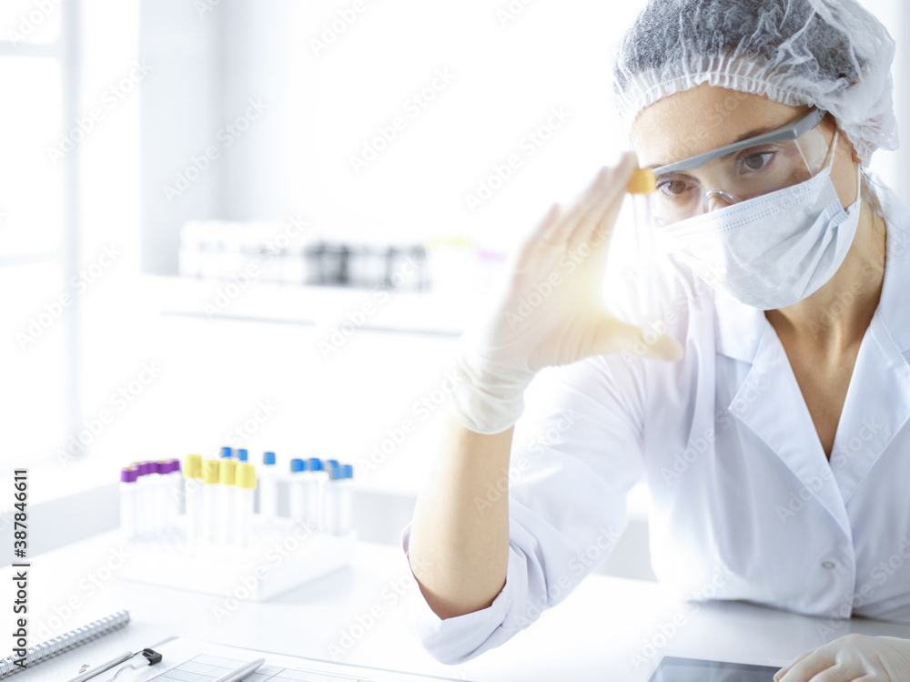 Naklejka premium Professional female scientist in protective eyeglasses researching tube with reagents in sunny laboratory. Medicine and science researching