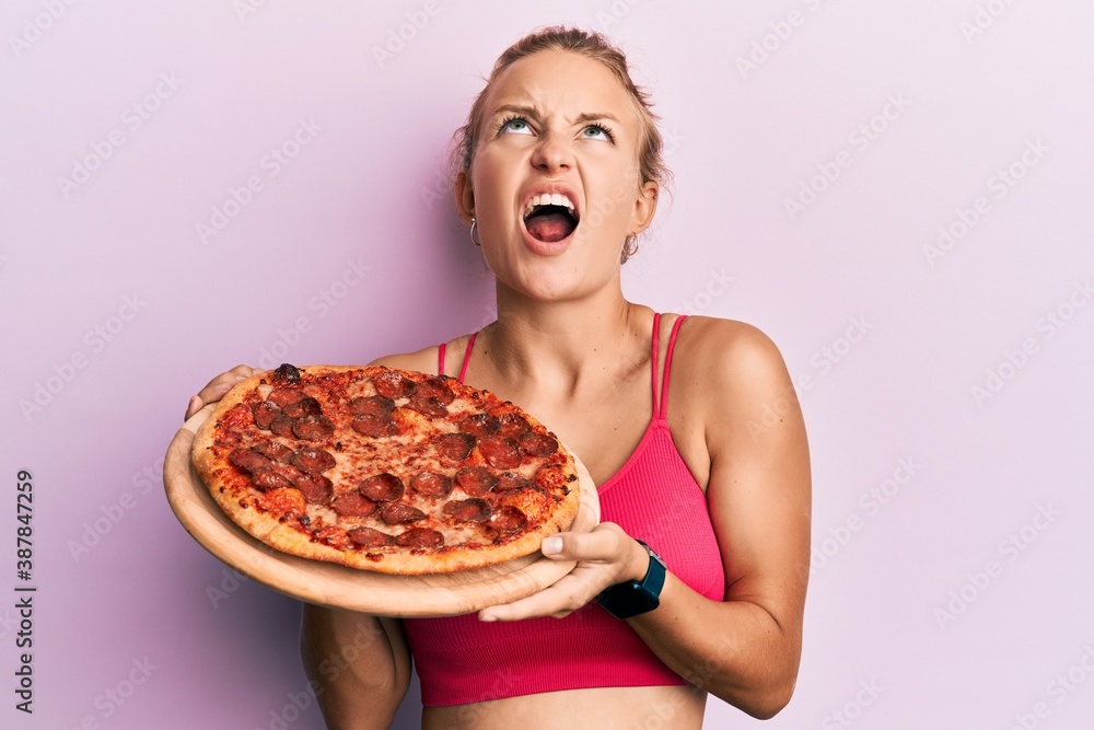 Beautiful caucasian woman holding italian pizza angry and mad screaming frustrated and furious, shouting with anger looking up. Beautiful caucasian woman holding italian pizza angry and mad screaming frustrated and furious, shouting with anger looking up.